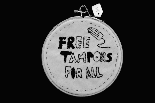 Greyscale. An embroidery hoop with the phrase "Free Tampons For All" stitched onto cloth in all-caps. A tampon is stitched into the top-right of the cloth, and there is a stitched border.