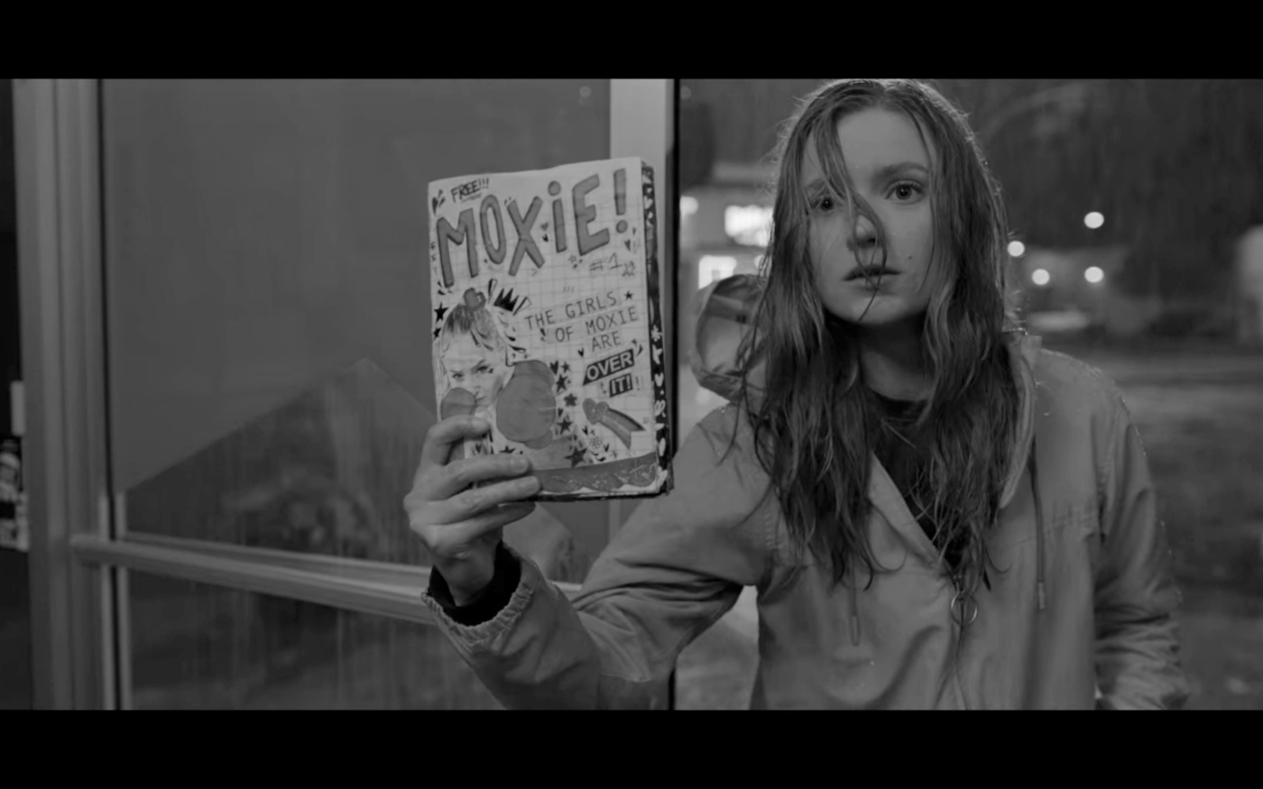 Greyscale. The film's protagonist, 16-year-old Vivian, holds up a copy of the Moxie zine. A white girl, with long blonde hair, she looks earnest and naive - though the graphic design on the front cover is bold. The free zine shows a white girl with boxing gloves on with the headline, "The Girls Of Moxie Are Over It!"
