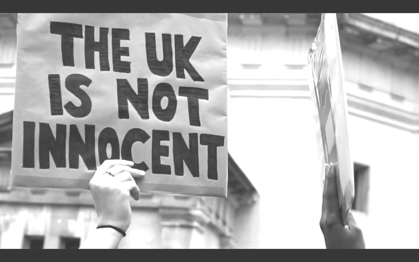 Still from Alberta Whittle’s film ‘Holding the line’. A white hand holds up a sign saying “The UK is not innocent”. Next to them a black hand is holding a sign sideways that is not readable from that angle.